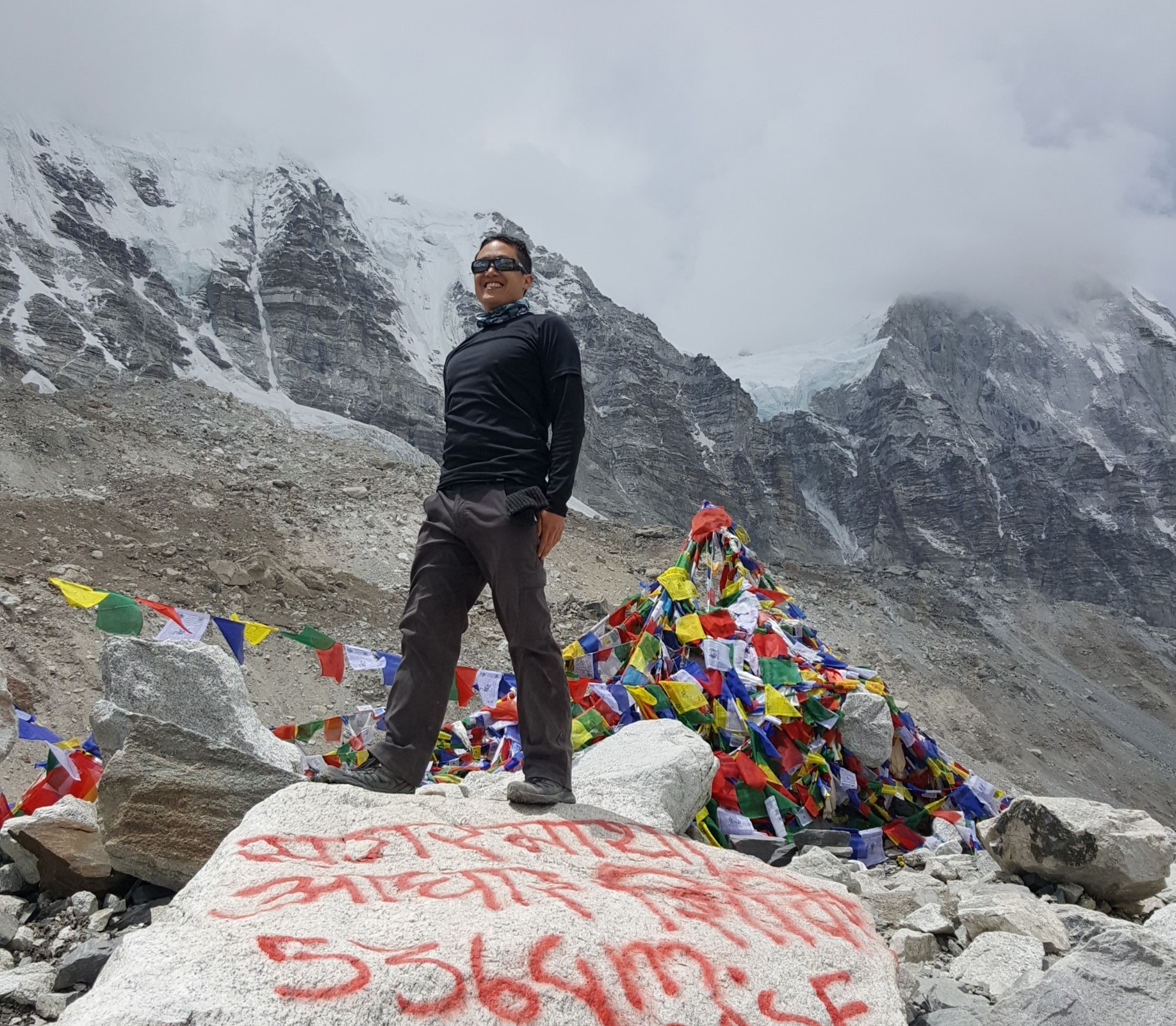 Edward standing on a boulder at Everest Base Camp, surrounded by prayer flags and snowy peaks